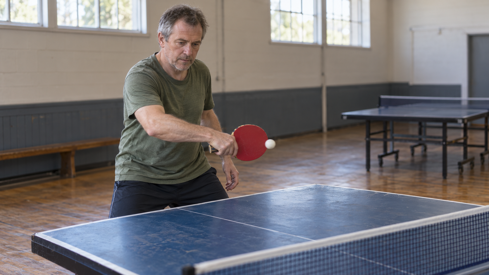 A recreational table-tennis player practices a backhand return with one paddle in a community hall