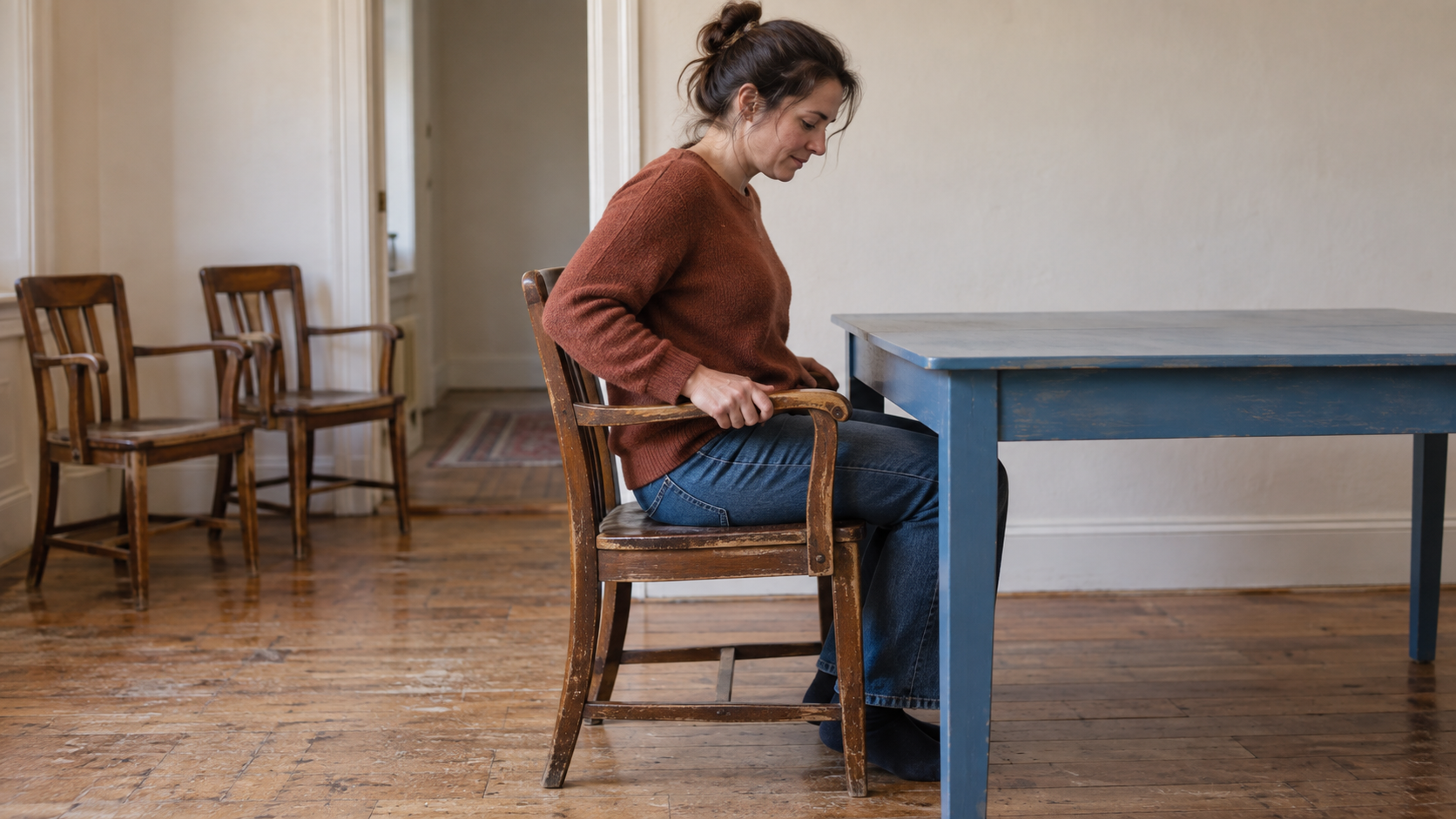 A woman checks whether a worn wooden armchair fits comfortably at a blue table while two matching chairs wait nearby.