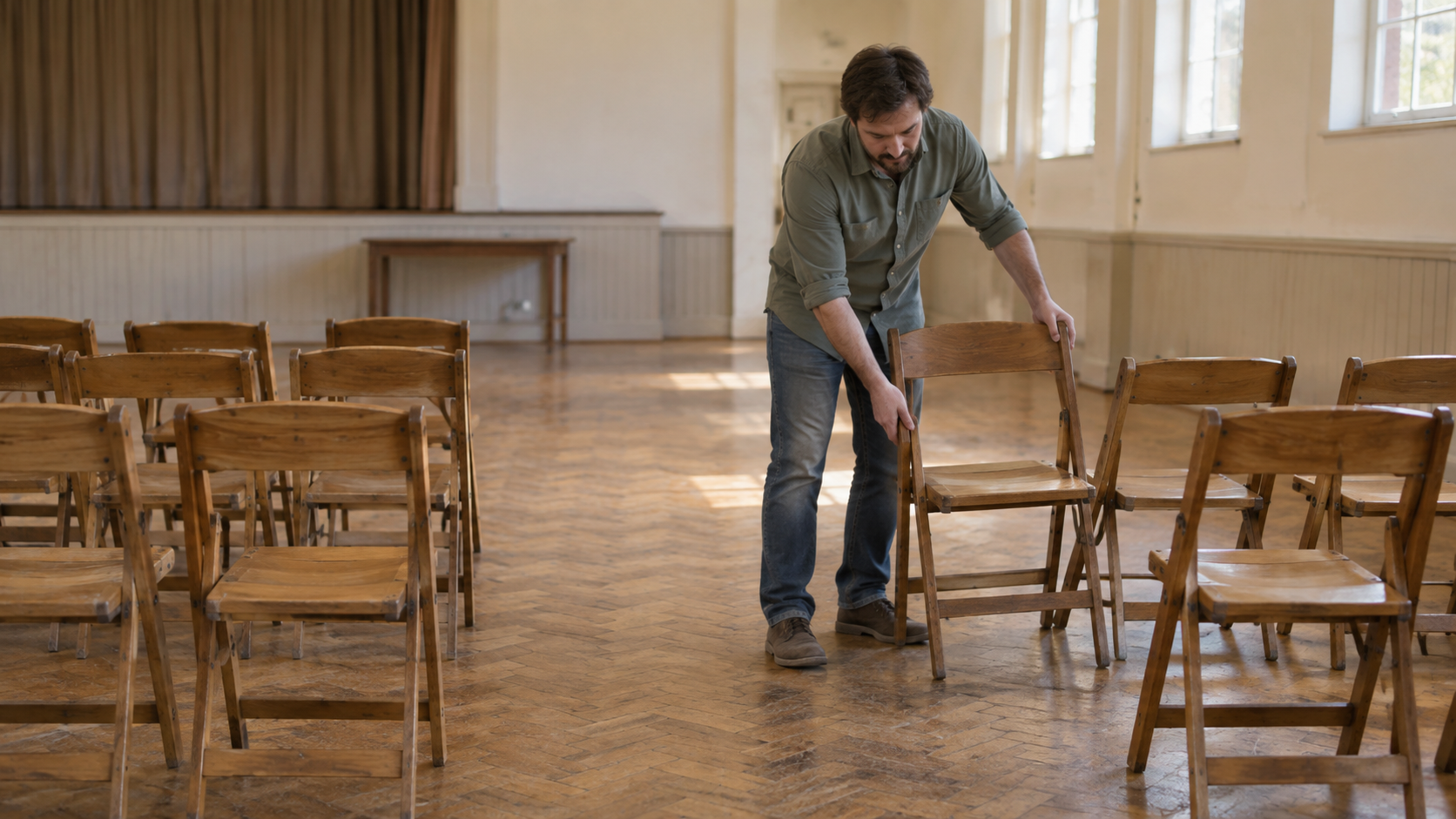 A community-hall volunteer moving a wooden folding chair beside a row to clear the central aisle